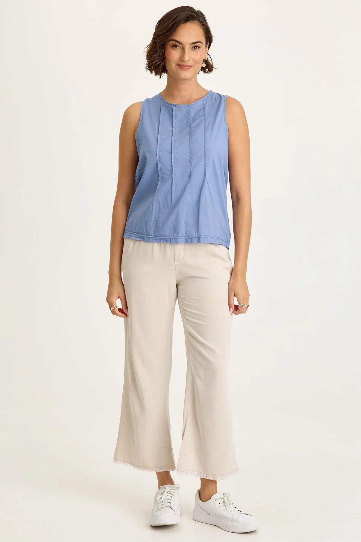 Full body view of a woman wearing a blue sleeveless top and beige pants on a white background