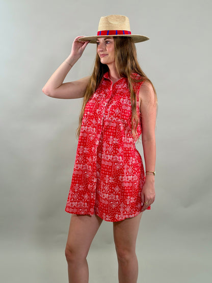 Woman looking to the side wearing a red sleeveless romper and a straw hat with a colorful band against a plain background