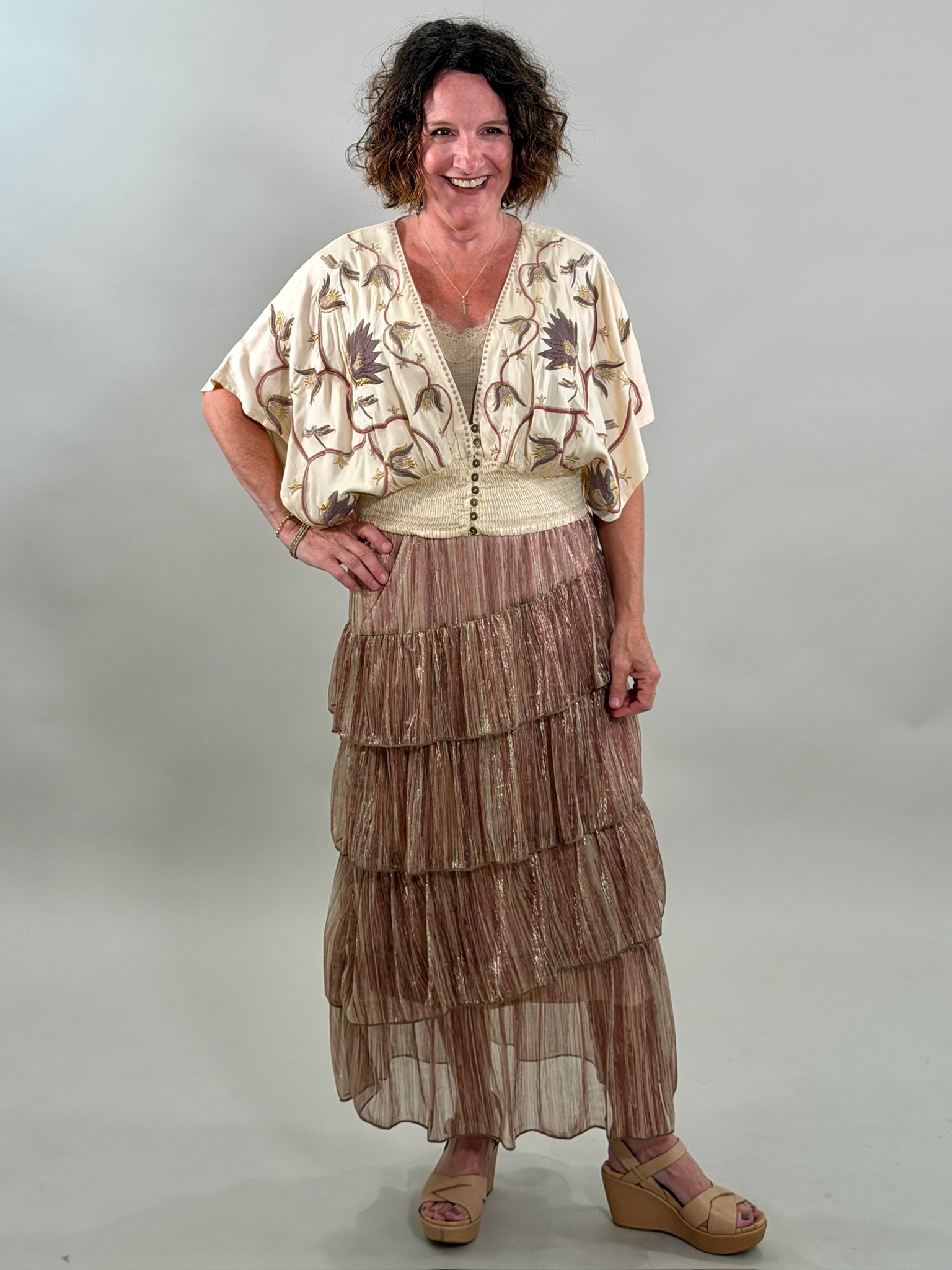 A smiling woman with her hand on her hip wearing a cream-colored bohemian blouse with purple and gold floral embroidery, a smocked waist, and a tan lace bralette underneath.