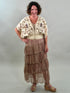 A smiling woman with her hand on her hip wearing a cream-colored bohemian blouse with purple and gold floral embroidery, a smocked waist, and a tan lace bralette underneath.