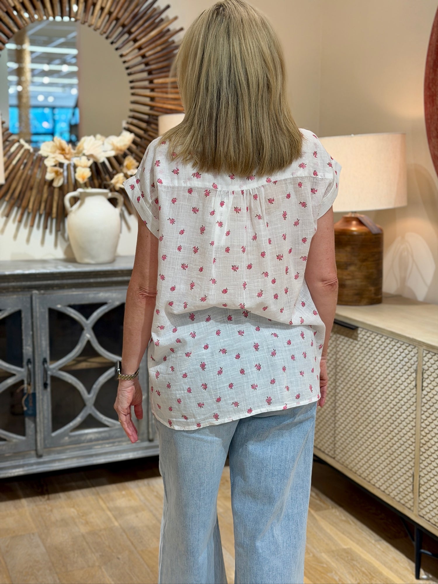 Rear view of a blonde woman wearing a white short-sleeve cotton button-up with a pink floral print and light-wash wide-leg jeans.