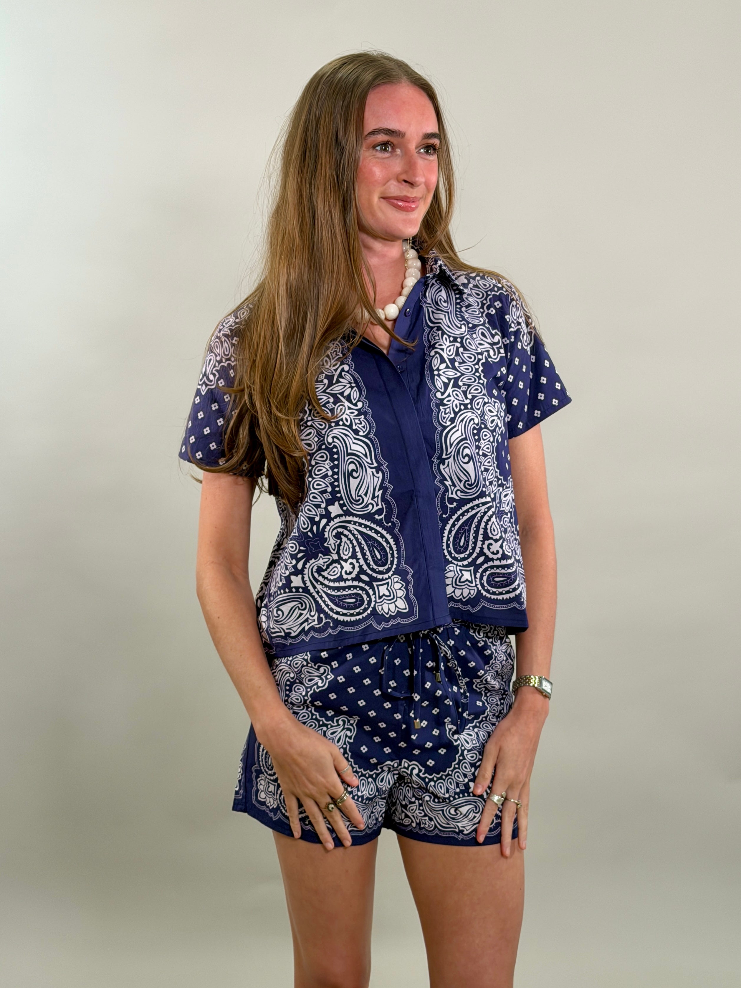A smiling woman with long brown hair looks to her side while wearing a navy blue and white paisley bandana-print short-sleeve button-down shirt paired with matching printed drawstring shorts.