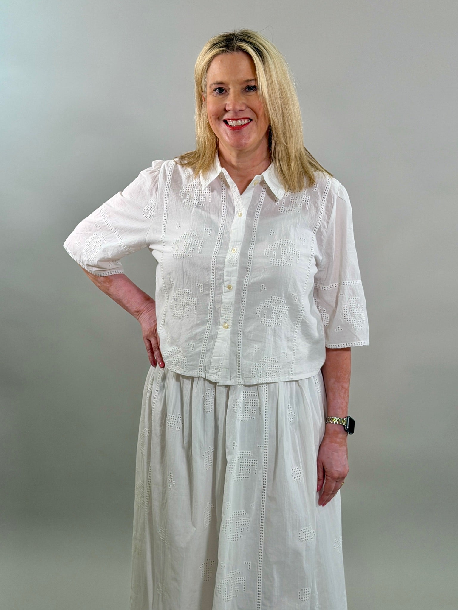 A smiling blonde woman with her hand on her hip wearing a white geometric eyelet cropped button-up shirt with a matching maxi skirt.