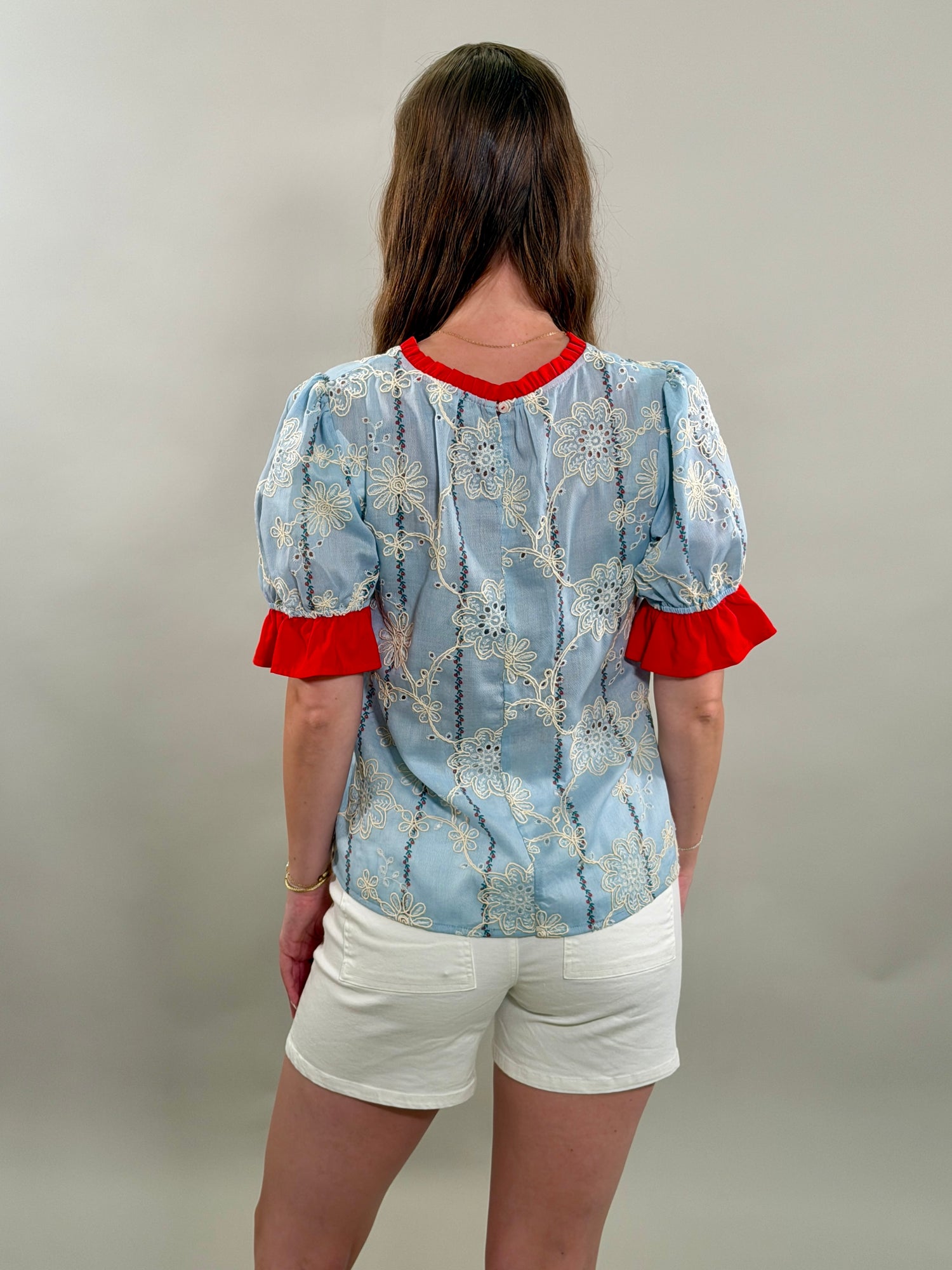 A rear view of a woman with long brown hair stands with her hand in her pocket, wearing a light blue cotton blouse with white floral embroidery and red ruffled sleeves.