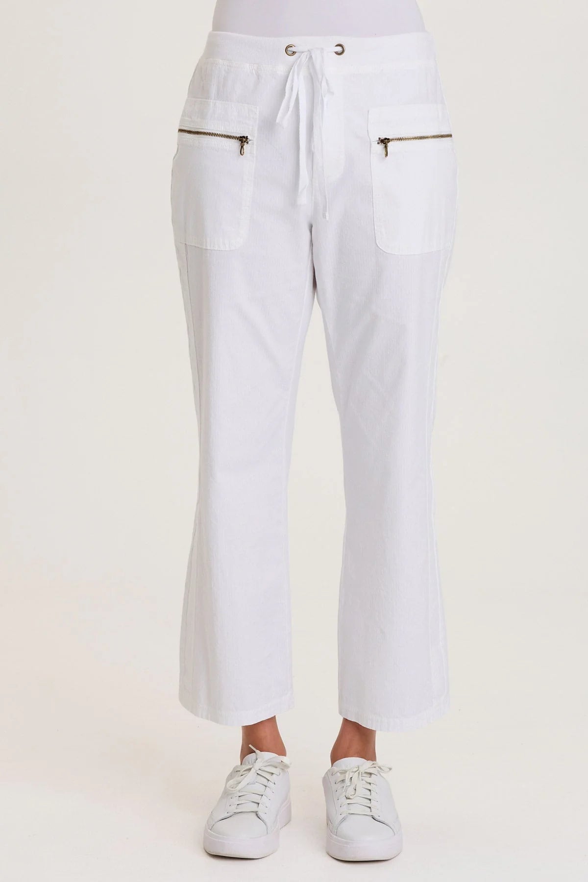 Front view of a woman wearing white structured seersucker flared pants with a drawstring waist and brass zipper pocket accents, paired with white sneakers.