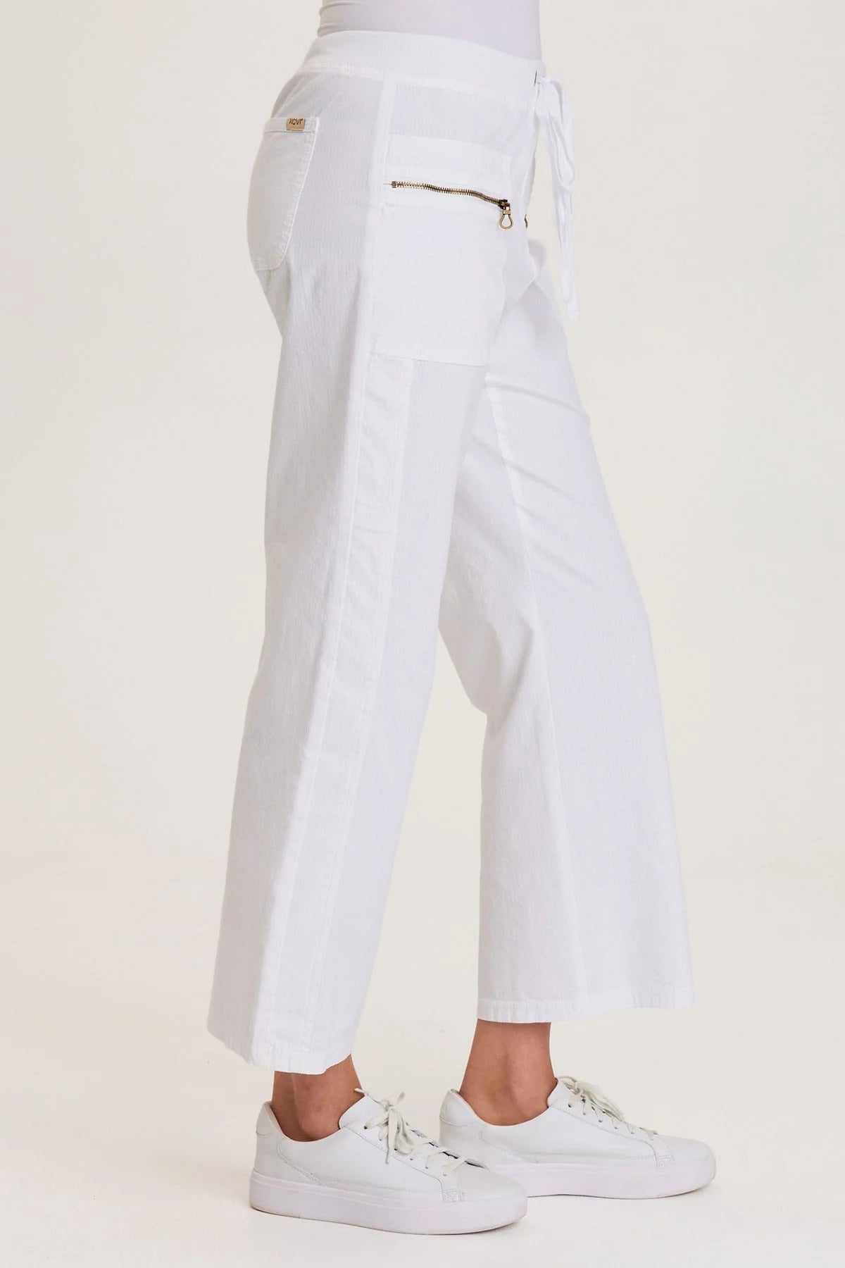 Side view of a woman wearing white structured seersucker flared pants with a drawstring waist and brass zipper pocket accents, paired with white sneakers.