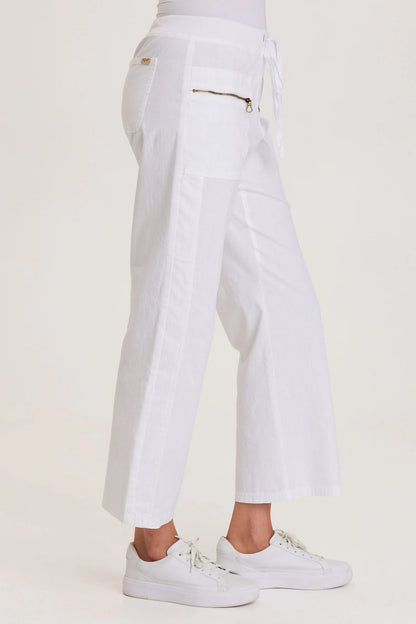 Side view of a woman wearing white structured seersucker flared pants with a drawstring waist and brass zipper pocket accents, paired with white sneakers.
