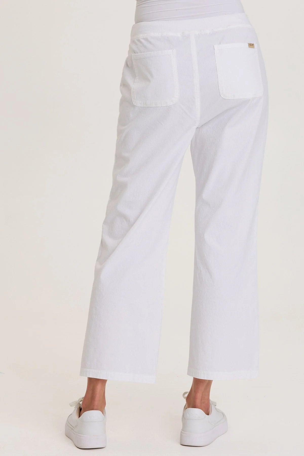 Rear view of a woman wearing white structured seersucker flared pants with a drawstring waist and brass zipper pocket accents, paired with white sneakers.