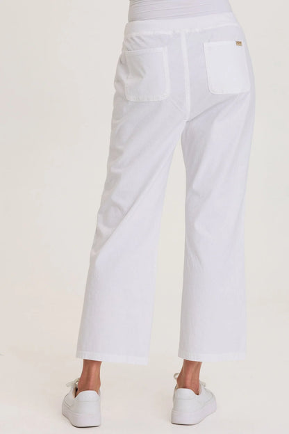 Rear view of a woman wearing white structured seersucker flared pants with a drawstring waist and brass zipper pocket accents, paired with white sneakers.