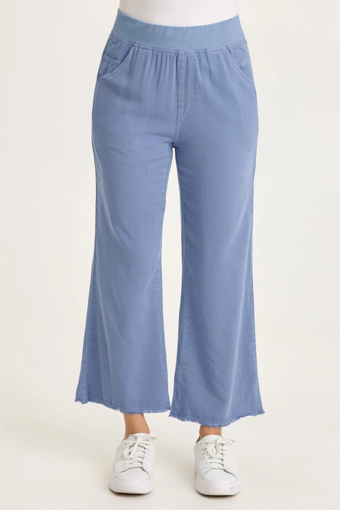 Close-up of a woman wearing light blue wide-leg twill pants with front pockets and a raw-edge frayed hem, paired with white sneakers.