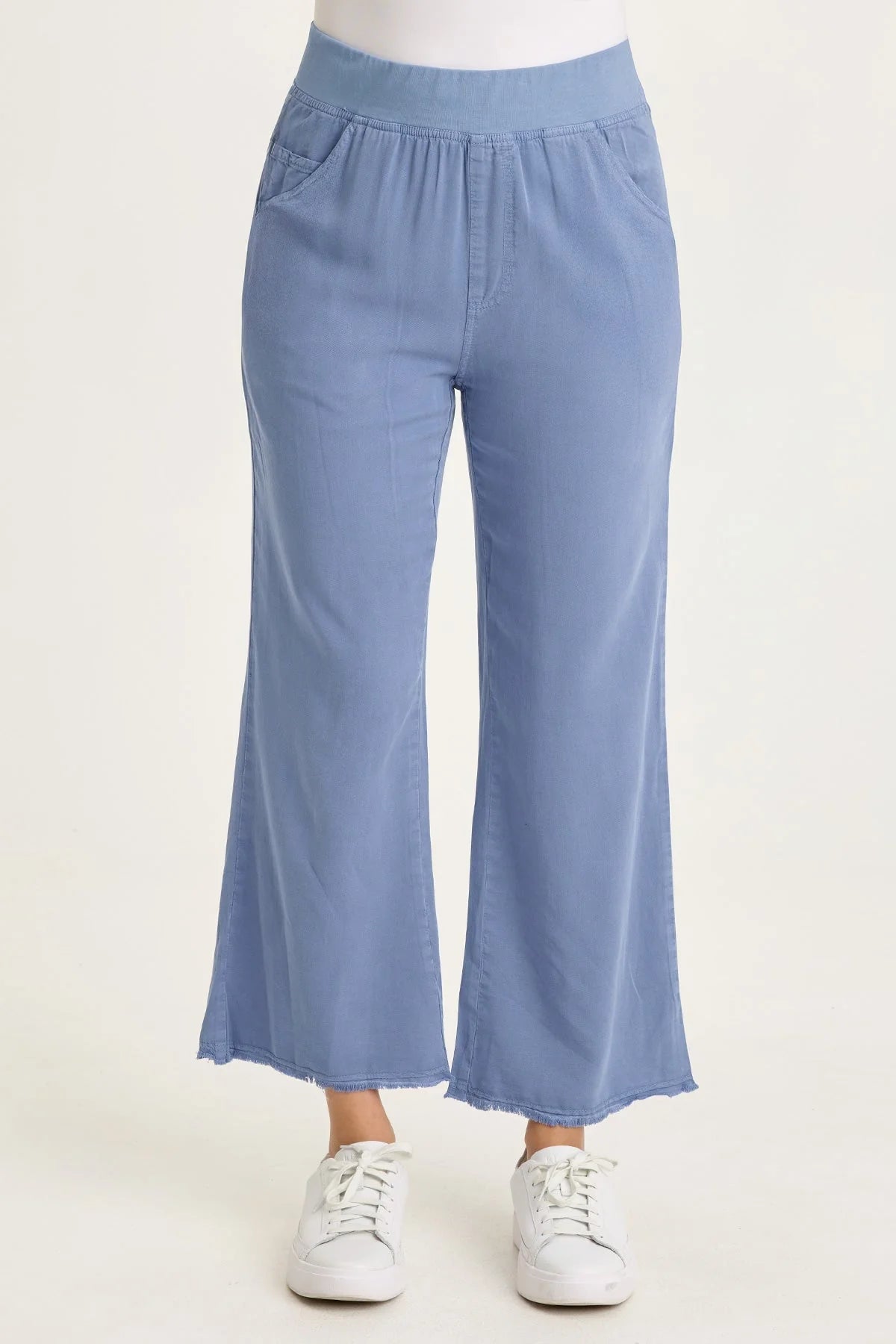 Close-up of a woman wearing light blue wide-leg twill pants with front pockets and a raw-edge frayed hem, paired with white sneakers.
