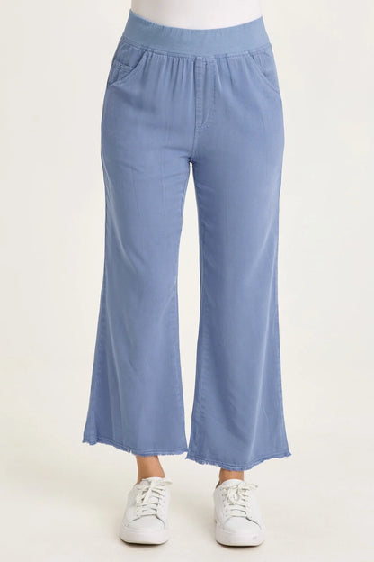 Close-up of a woman wearing light blue wide-leg twill pants with front pockets and a raw-edge frayed hem, paired with white sneakers.