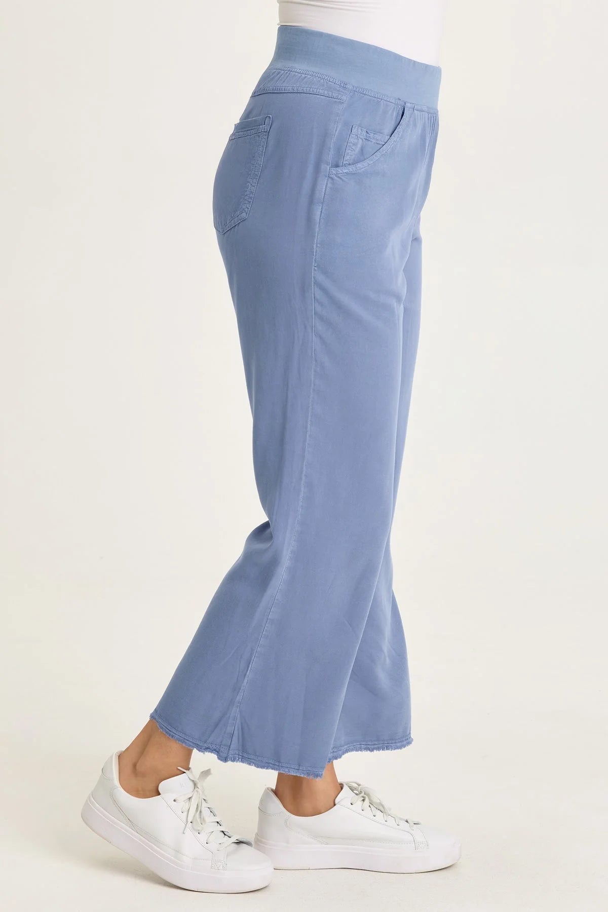 Side view of a woman wearing light blue wide-leg twill pants with front pockets and a raw-edge frayed hem, paired with white sneakers.