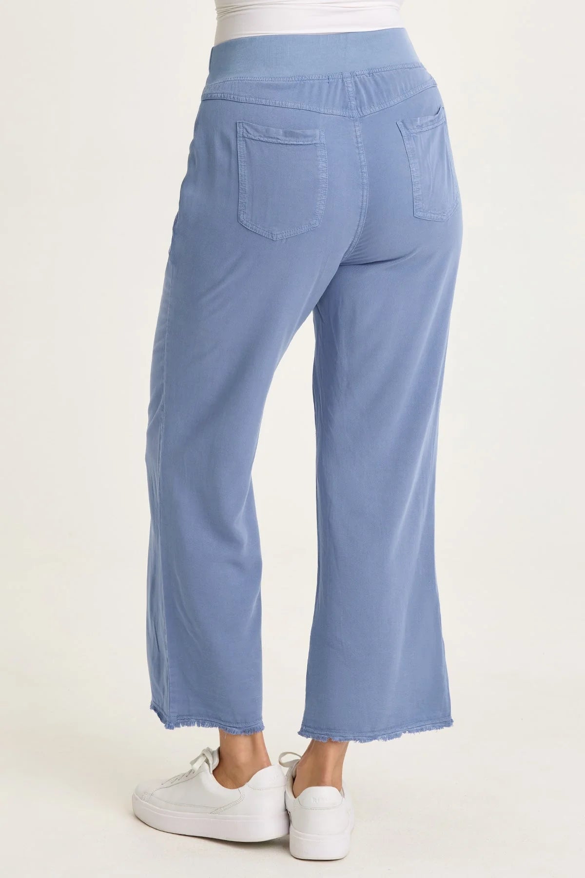 Rear view of a woman wearing light blue wide-leg twill pants with front pockets and a raw-edge frayed hem, paired with white sneakers.