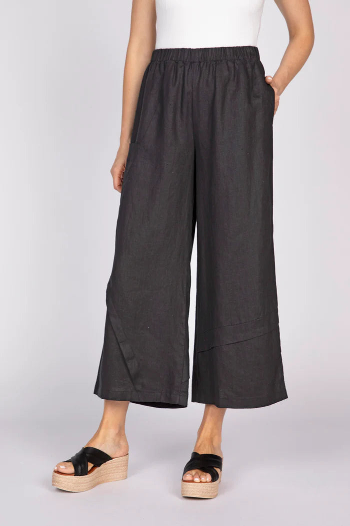 A front view of a woman with a hand in her pocket wearing black wide-leg linen pants with unique pleat details at the hem and black platform sandals.