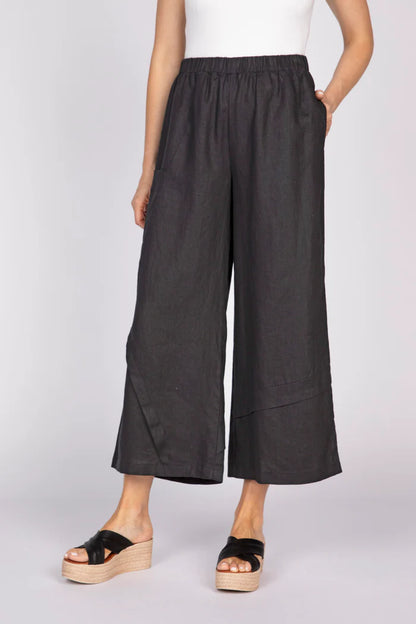 A front view of a woman with a hand in her pocket wearing black wide-leg linen pants with unique pleat details at the hem and black platform sandals.