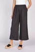 A front view of a woman with a hand in her pocket wearing black wide-leg linen pants with unique pleat details at the hem and black platform sandals.