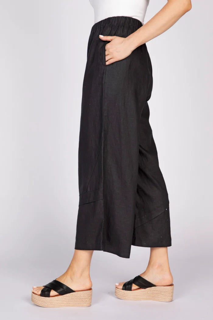 A side view of a woman with a hand in her pocket wearing black wide-leg linen pants with unique pleat details at the hem and black platform sandals.