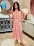 A woman with dark hair smiling while wearing a pink and orange patterned column maxi dress with a white braided rope belt and tan platform mules.