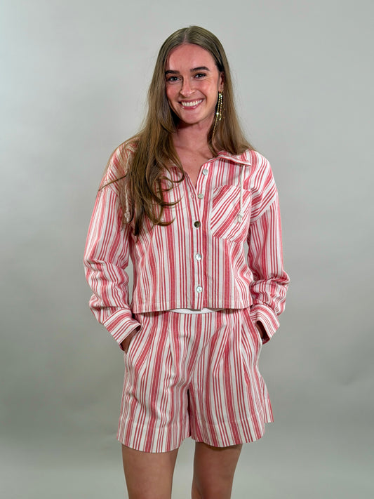 A smiling woman with long brown hair stands with her hands in her pockets, wearing a relaxed matching set featuring a red and white candy-striped cropped long-sleeve button-down shirt and matching pleated shorts.