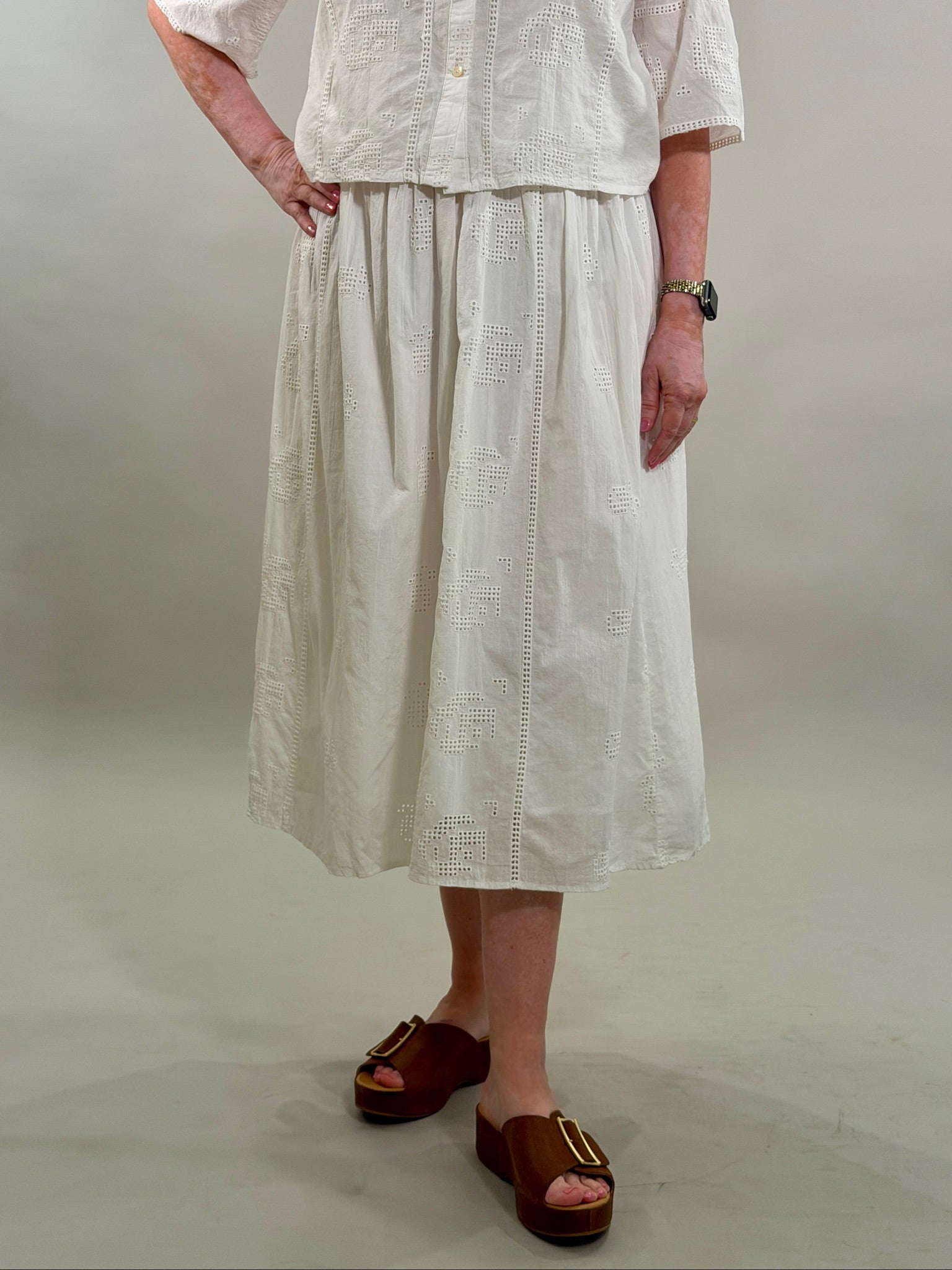Close-up of a woman wearing a white geometric eyelet midi skirt paired with brown platform slide sandals.