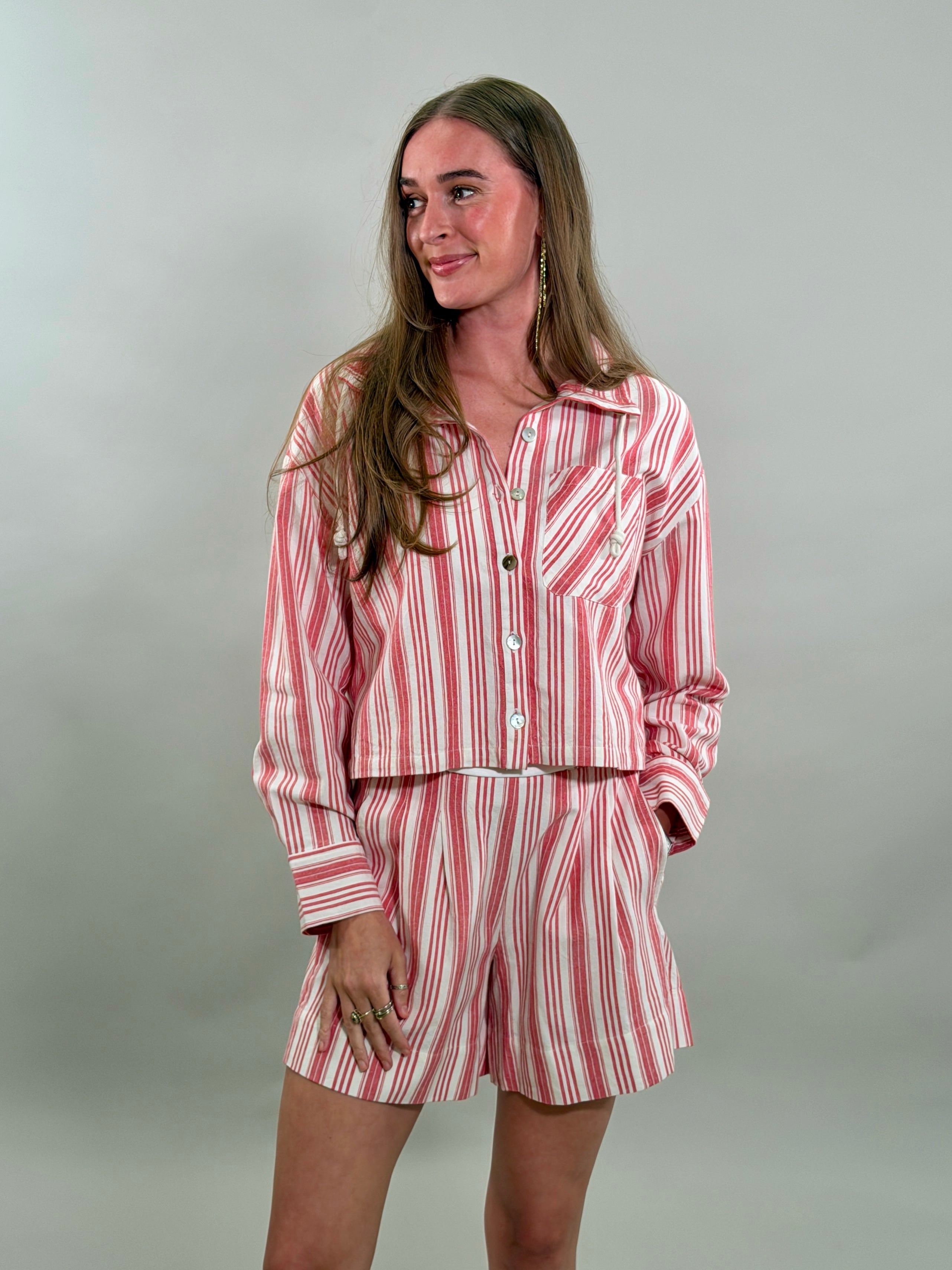 A smiling woman with long brown hair stands with her hand in her pockets, wearing a relaxed matching set featuring a red and white candy-striped cropped long-sleeve button-down shirt and matching pleated shorts.