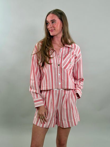 A smiling woman with long brown hair stands with her hand in her pockets, wearing a relaxed matching set featuring a red and white candy-striped cropped long-sleeve button-down shirt and matching pleated shorts.
