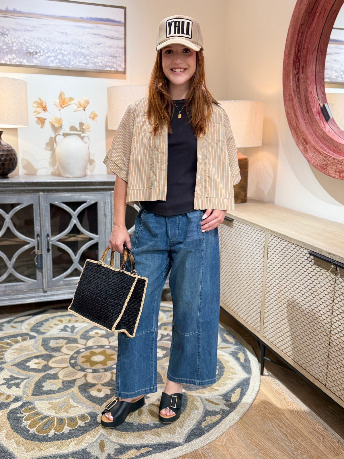 A woman stands in a styled indoor room wearing a beige cropped jacket, black top, wide-leg blue jeans, black slide sandals, and a beige cap with &