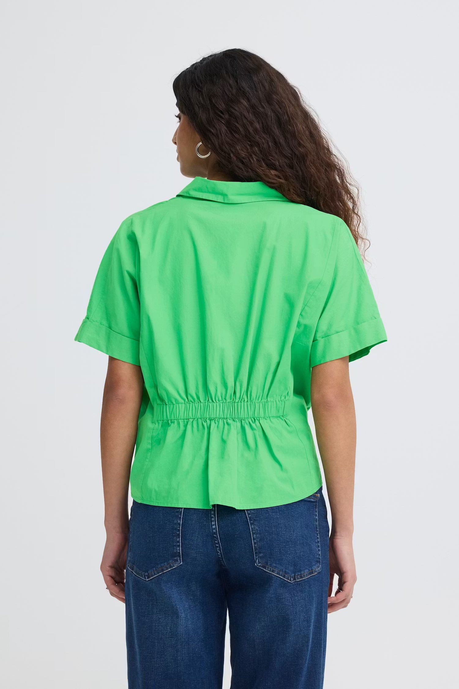 Rear view of a woman wearing a bright green, boxy short sleeve cotton shirt with cinched back with dark wash jeans.