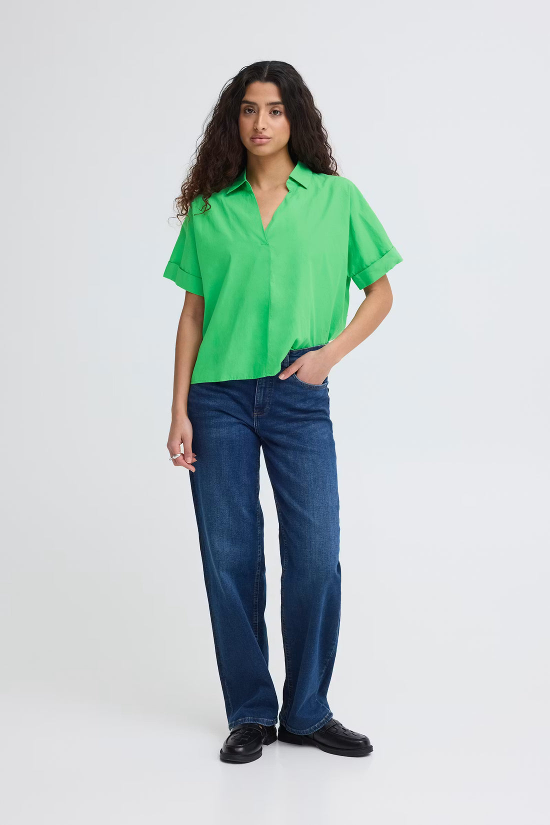 Full body view of a woman wearing a bright green, boxy short sleeve cotton shirt with an open collar and V-neck, paired with blue jeans.