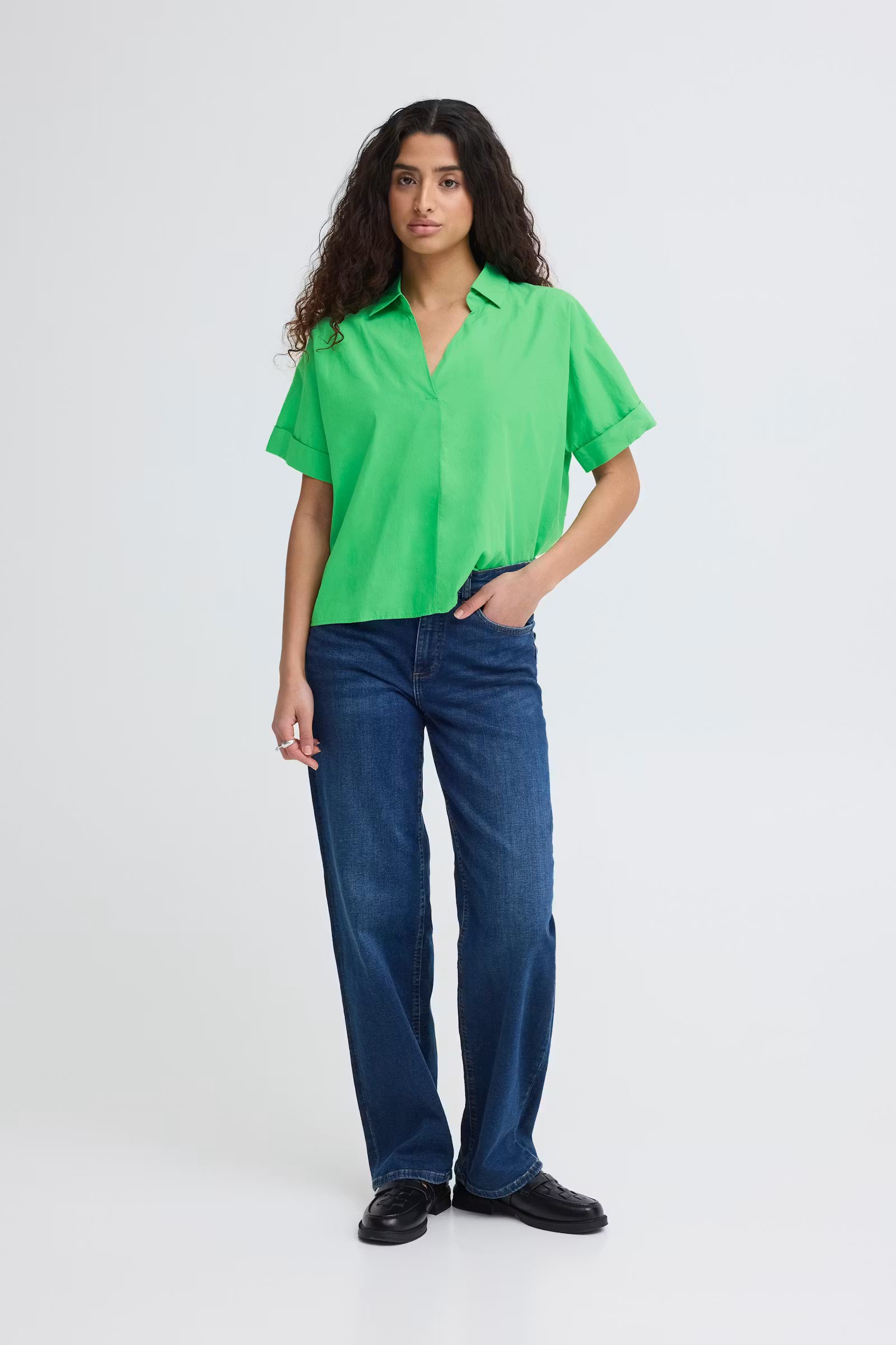 Full body view of a woman wearing a bright green, boxy short sleeve cotton shirt with an open collar and V-neck, paired with blue jeans.