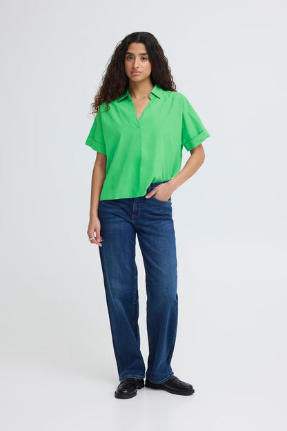 Full body view of a woman wearing a bright green, boxy short sleeve cotton shirt with an open collar and V-neck, paired with blue jeans.