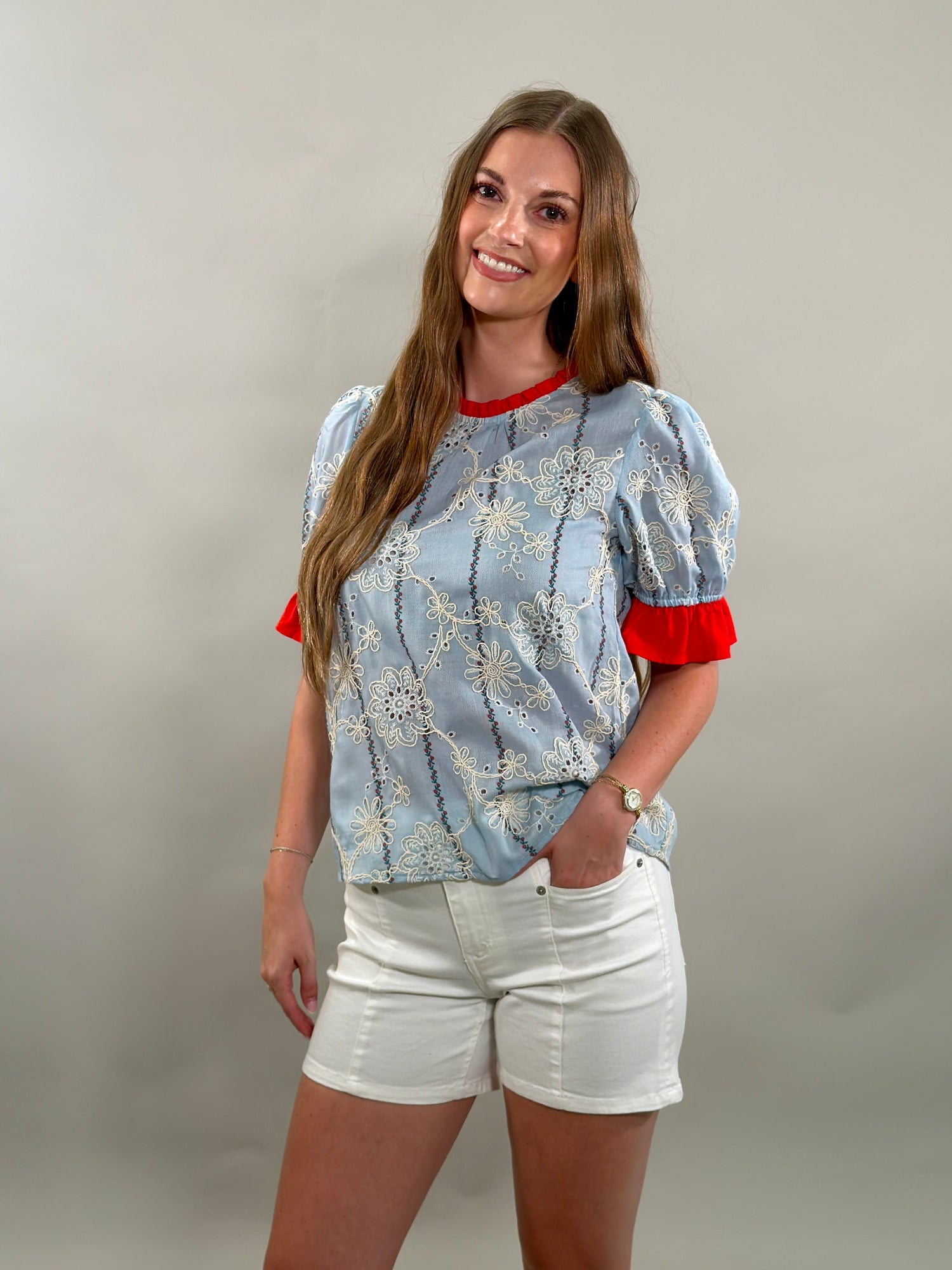 A smiling woman with long brown hair stands with her hand in her pocket, wearing a light blue cotton blouse with white floral embroidery and red ruffled sleeves.
