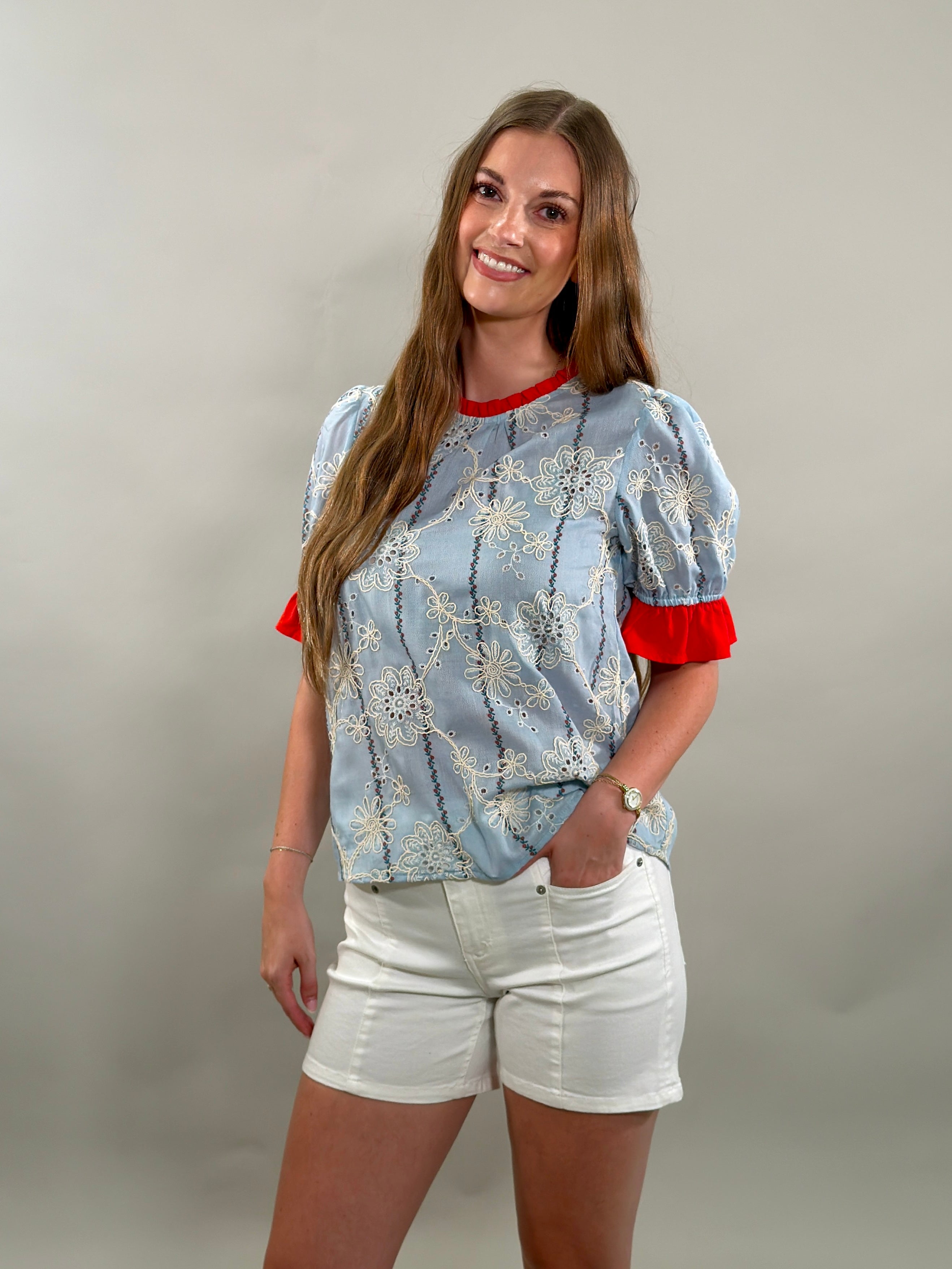 A smiling woman with long brown hair stands with her hand in her pocket, wearing a light blue cotton blouse with white floral embroidery and red ruffled sleeves.