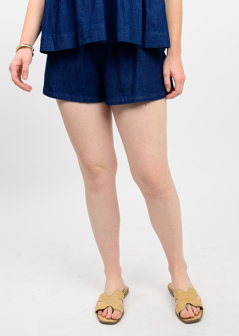 Woman wearing dark blue cotton shorts with a smocked waistband and woven slide sandals with hand on her hip.