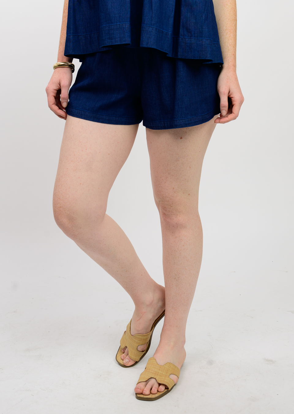 Woman wearing dark blue cotton shorts with a smocked waistband and woven slide sandals with a bent knee.