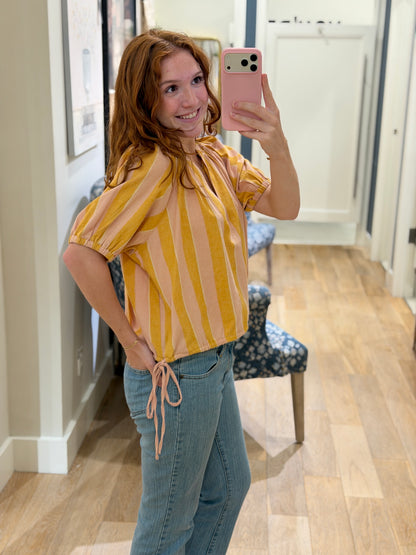 Side view of a smiling young woman taking a mirror selfie wearing a peach and mustard yellow striped cotton blouse with blue jeans.