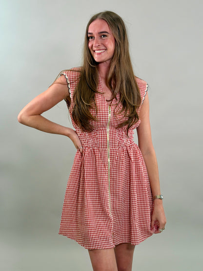 A smiling woman looking to the side with long brown hair wearing a red and white gingham sleeveless dress with a gold front zipper and a smocked waistband.