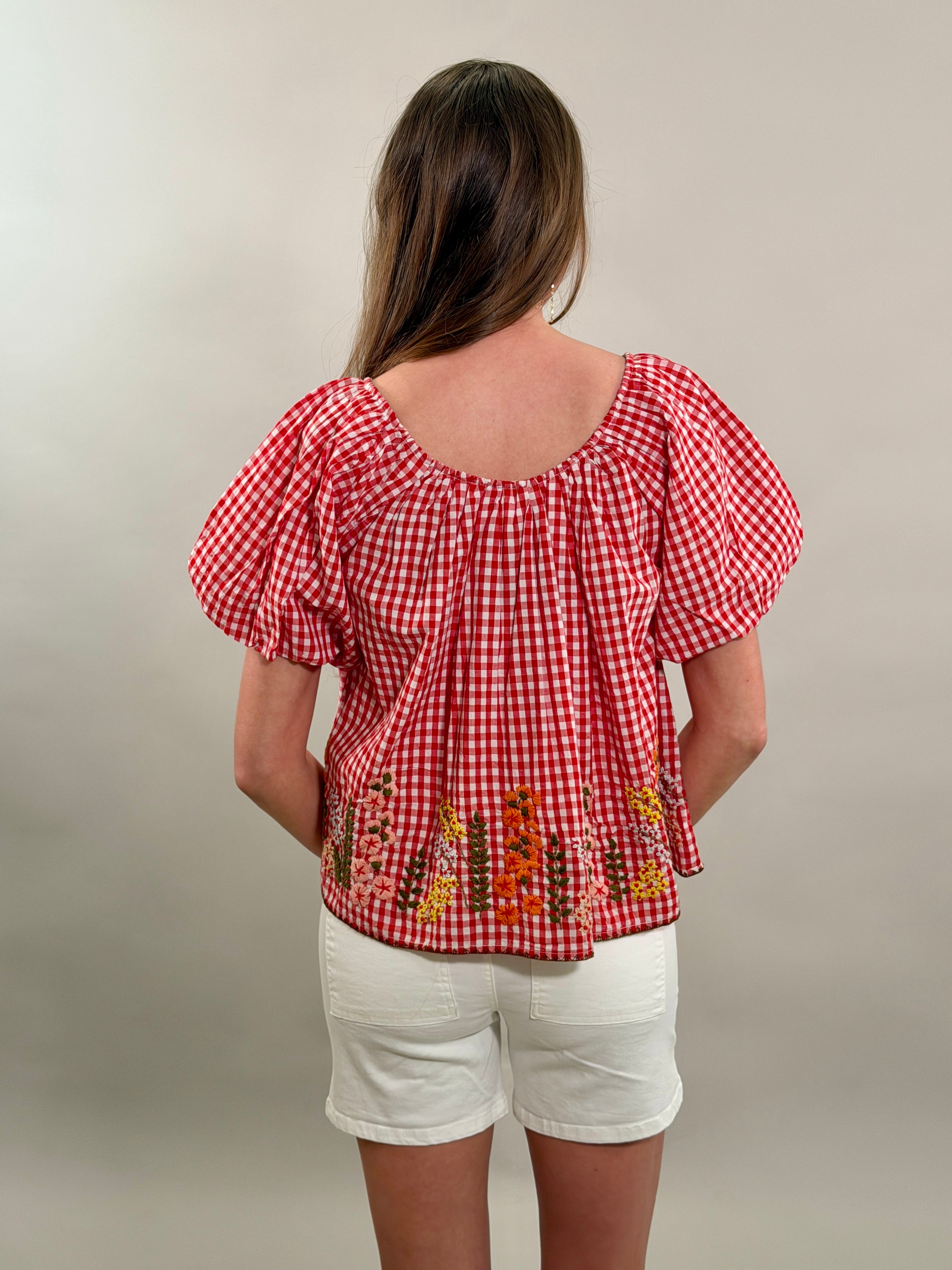 Rear view of a woman wearing a red checkered blouse with floral embroidery and white shorts on a plain background