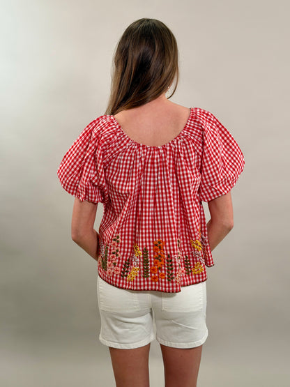 Rear view of a woman wearing a red checkered blouse with floral embroidery and white shorts on a plain background