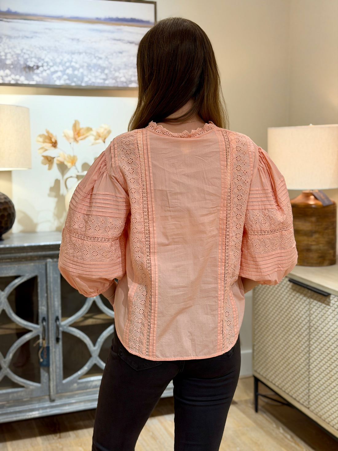 A woman viewed from behind wearing a peach-colored blouse with lace trim and puff sleeves, paired with black pants, standing in a home interior setting.