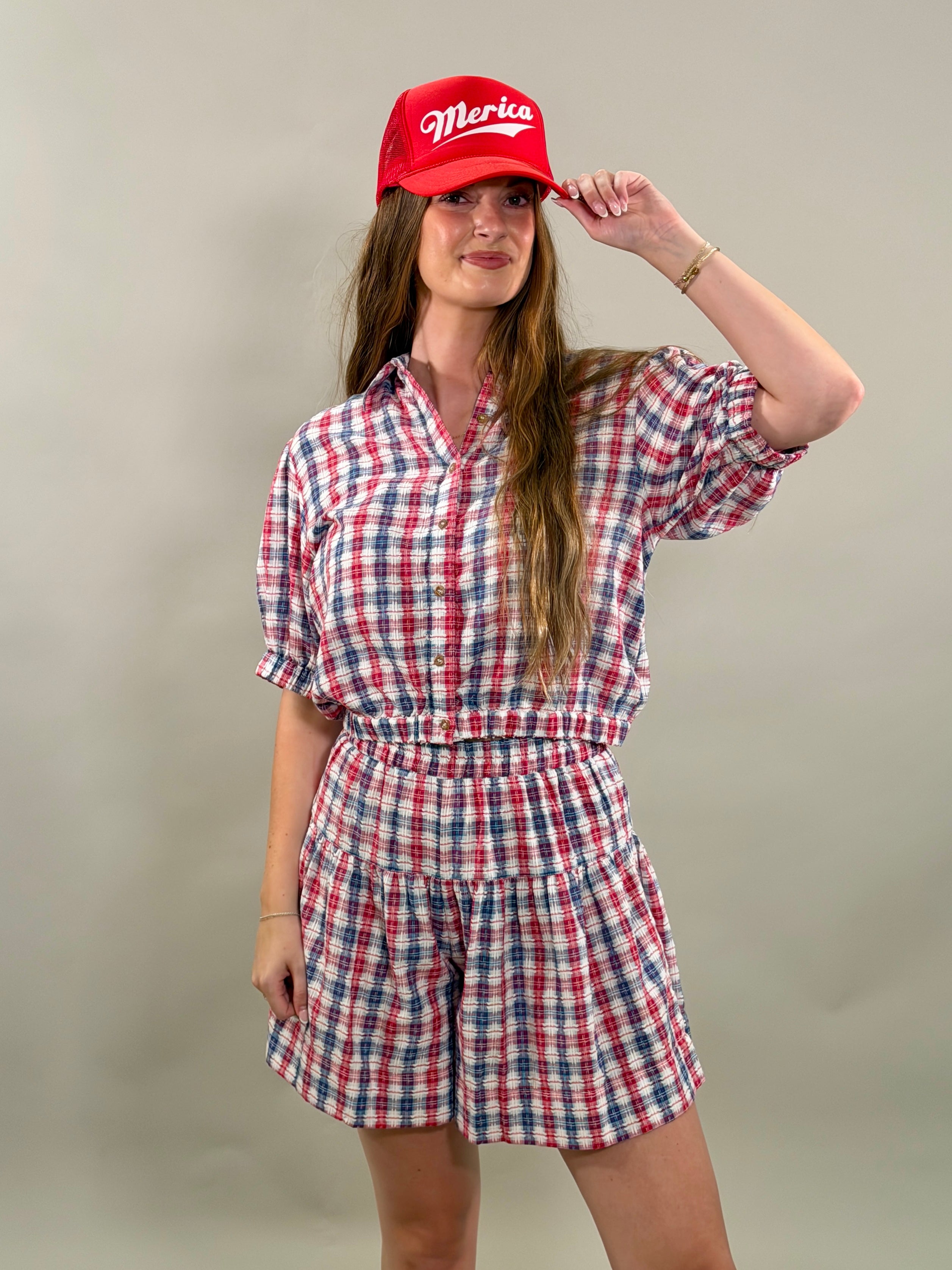 Woman smiling and wearing a red, navy, and white plaid short-sleeve cropped button-down shirt with a matching skort and a red baseball cap.