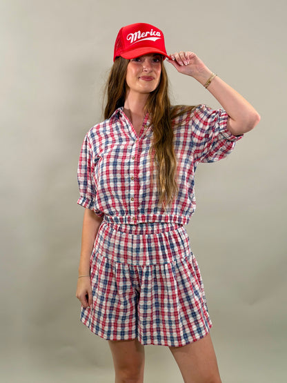 Woman smiling and wearing a red, navy, and white plaid short-sleeve cropped button-down shirt with a matching skort and a red baseball cap.