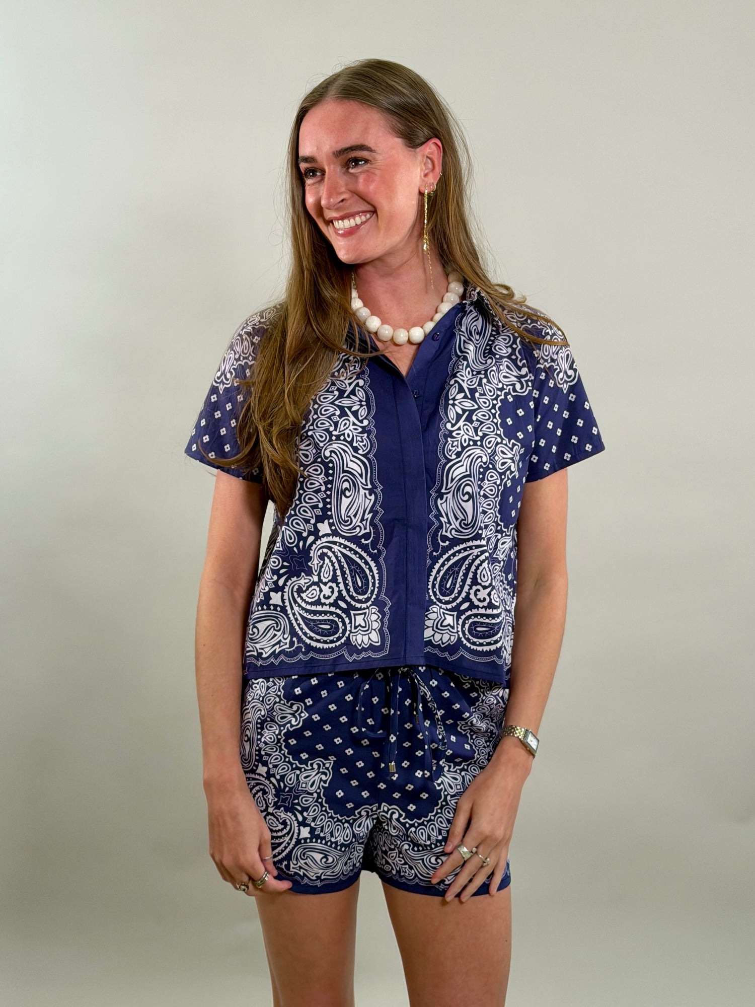 A smiling woman with long brown hair looks to her side while wearing a navy blue and white paisley bandana-print short-sleeve button-down shirt paired with matching printed drawstring shorts.