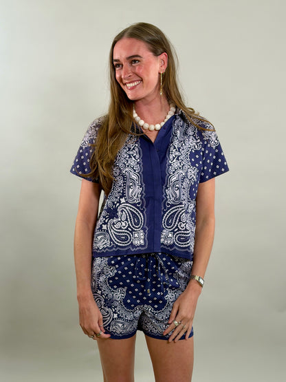 A smiling woman with long brown hair looks to her side while wearing a navy blue and white paisley bandana-print short-sleeve button-down shirt paired with matching printed drawstring shorts.