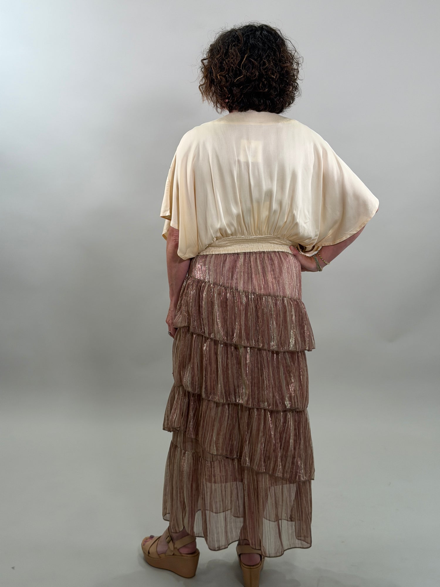 Rear view of a woman wearing a beige blouse and brown pleated skirt on a plain background