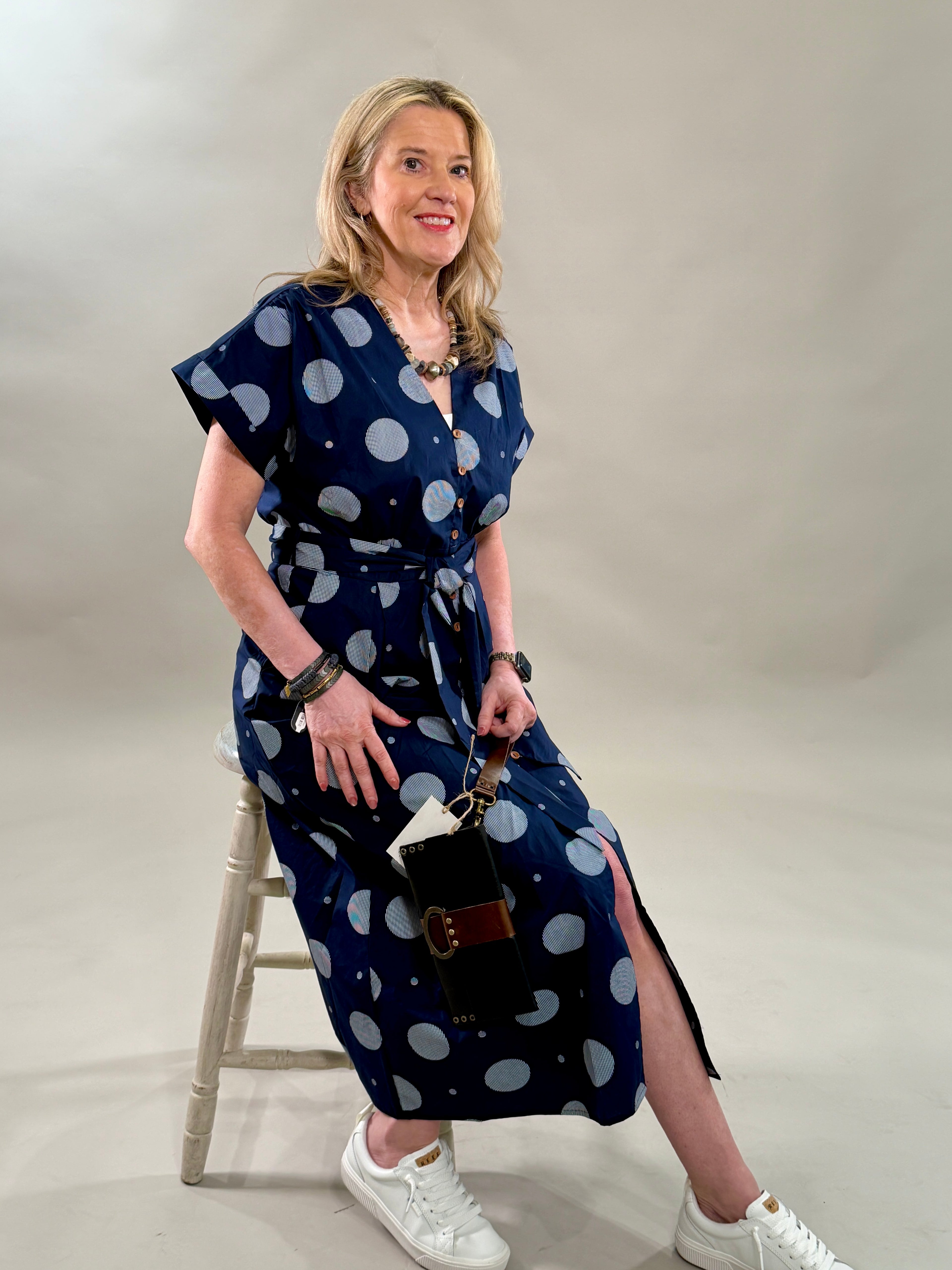 Woman sitting on a stool in a navy polka dot 100% cotton midi dress with a removable belt and white sneakers