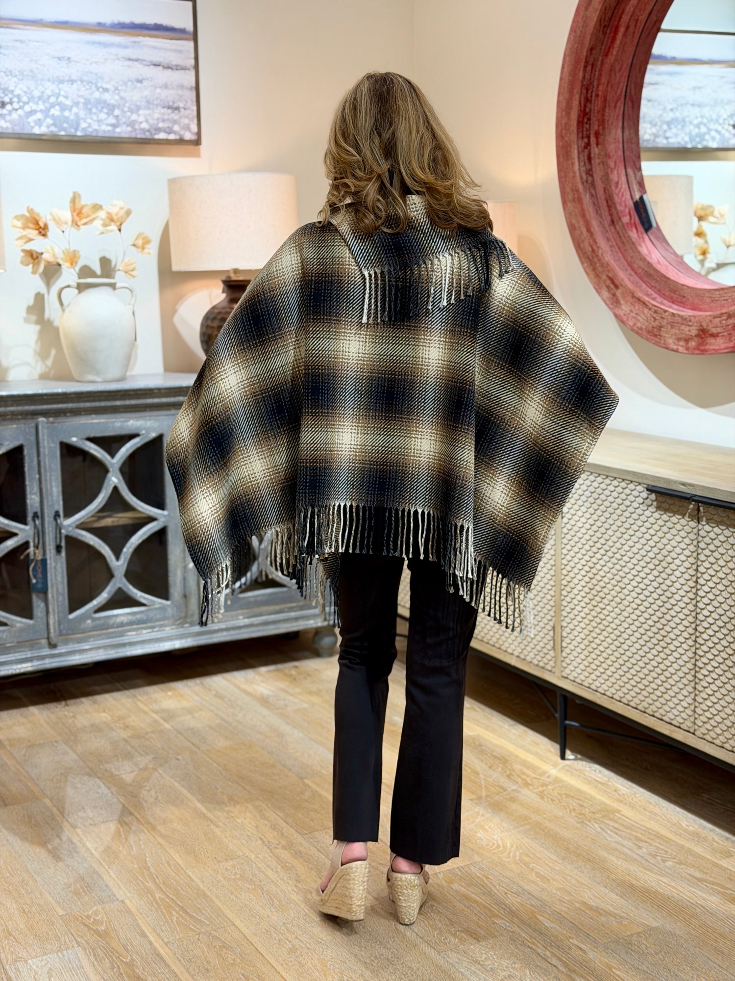Full body rear view of woman wearing a black, taupe, and mocha plaid poncho with a cowl neck and fringe trim.