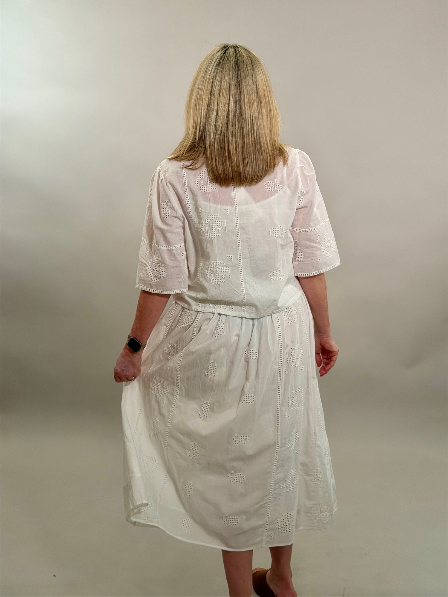 Rear view of a blonde woman wearing a white geometric eyelet cropped button-up shirt with a matching maxi skirt.