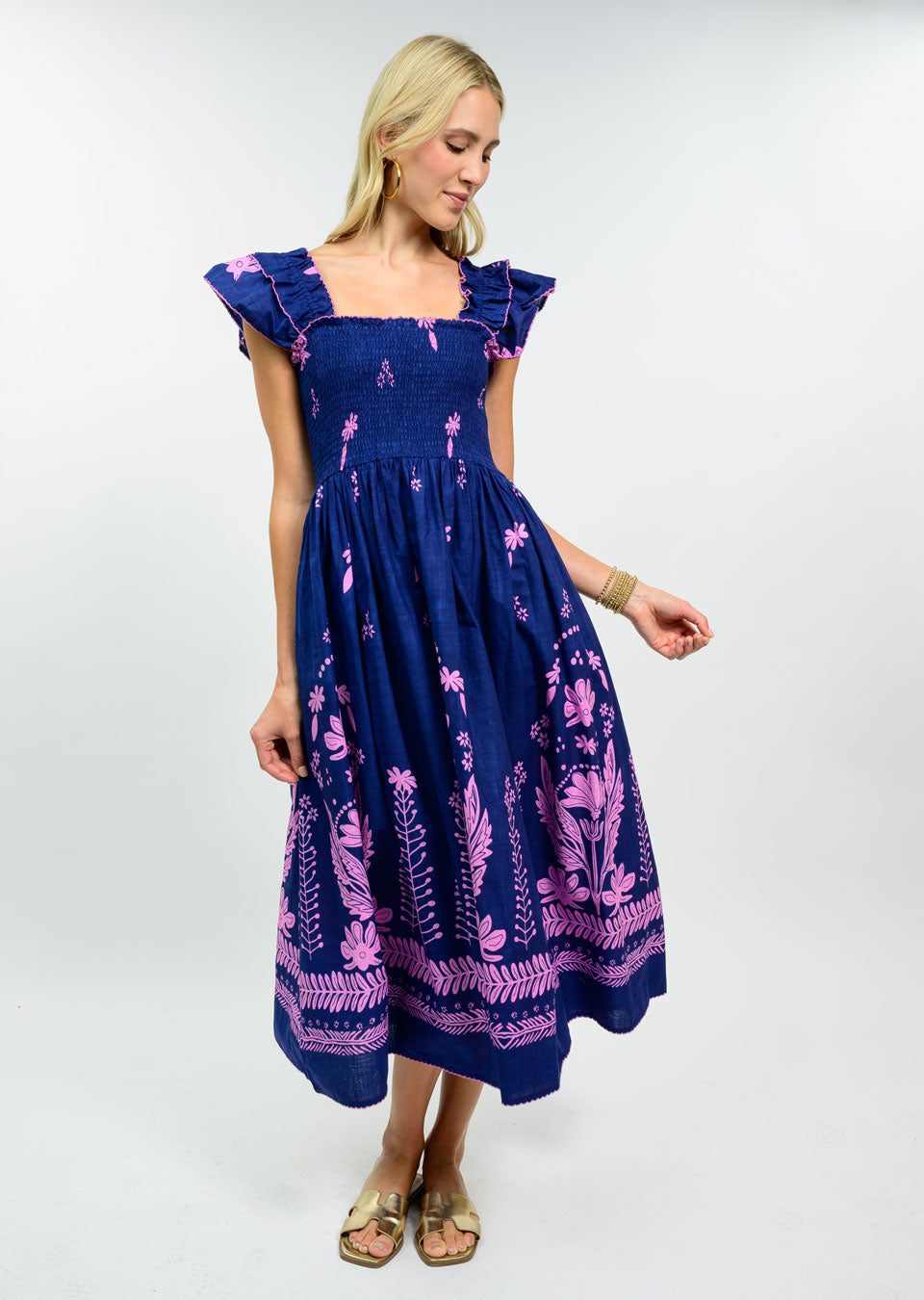 Woman wearing a navy blue 100% cotton midi dress with a smocked bodice, pink floral embroidery, and ruffle cap sleeves.